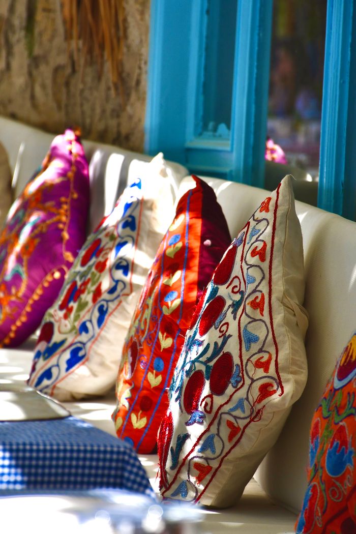 services-04 Brightly colored embroidered cushions on a sunlit bench with a rustic and cozy feel.