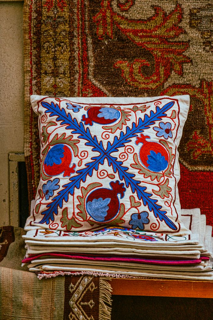 services-01 Vibrant embroidered pillow with blue and red patterns on stacked rustic rugs.