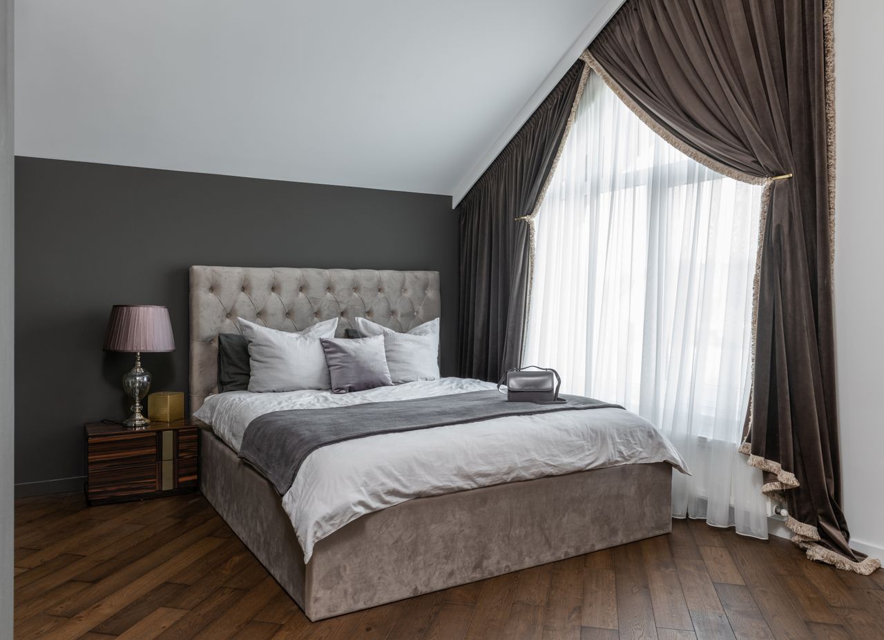 about-us Comfortable bed with blanket and cushions placed on parquet near window with curtains and lamp on bedside table in modern bedroom